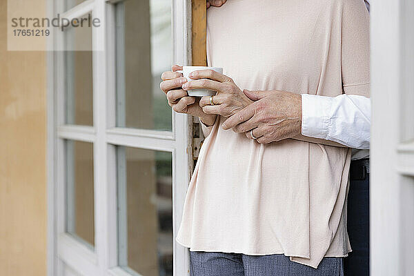 Ehemann hält die Hand seiner Frau und steht mit einer Kaffeetasse am Fenster