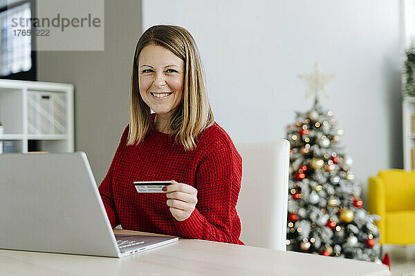 Glückliche Frau mit Kreditkarte und Laptop sitzt zu Hause am Tisch im Wohnzimmer