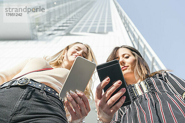 Lächelnde junge Frauen mit Mobiltelefon stehen vor dem Gebäude