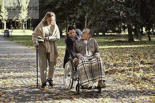 Behinderter älterer Mann unterhält sich mit Frau  die mit Stock geht  während Hausmeister Rollstuhl im Park schiebt