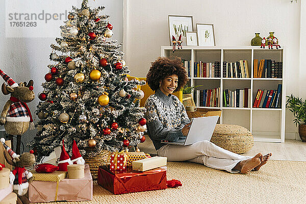 Glückliche junge Frau mit Laptop sitzt zu Hause am Weihnachtsbaum