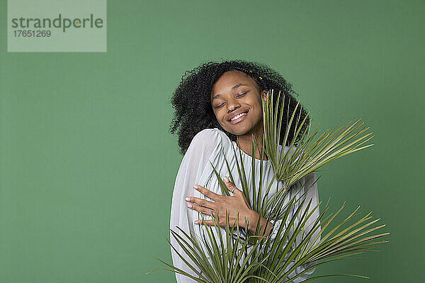 Glückliche junge Frau umarmt Palmzweige vor grünem Hintergrund
