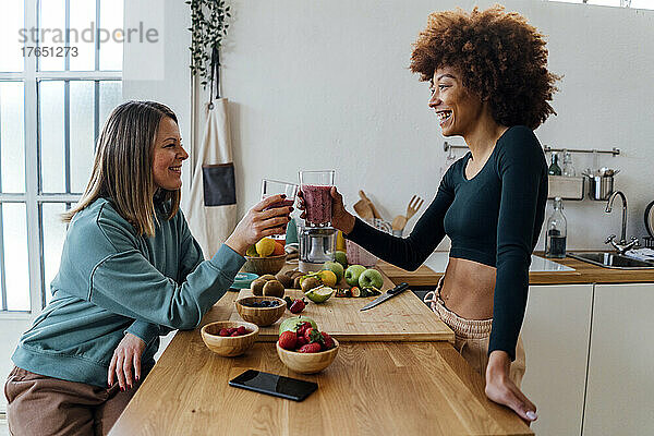 Glückliche junge Frau prostet mit einem Freund  der am Tisch in der Küche sitzt  ein Glas Smoothie an