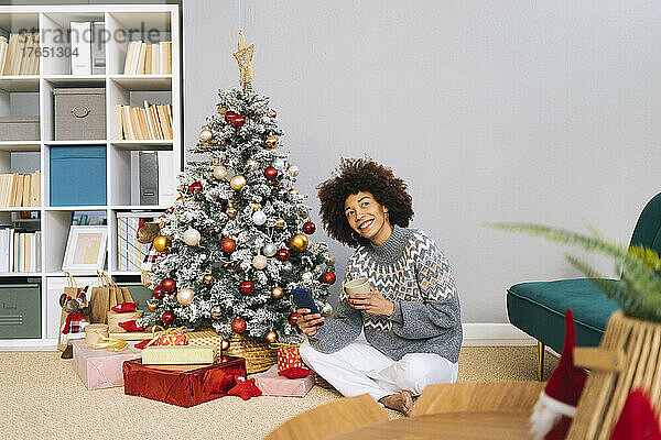 Lächelnde Frau sitzt zu Hause am Weihnachtsbaum und hält eine Tasse in der Hand