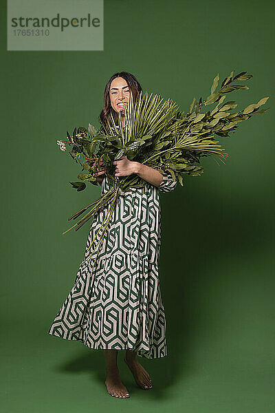 Smiling woman holding bunch of leaves standing against green background