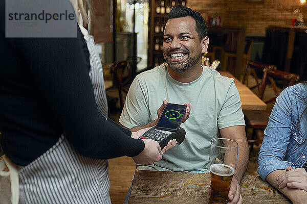 Lächelnder Mann  der in einer Bar mit dem Smartphone bezahlt