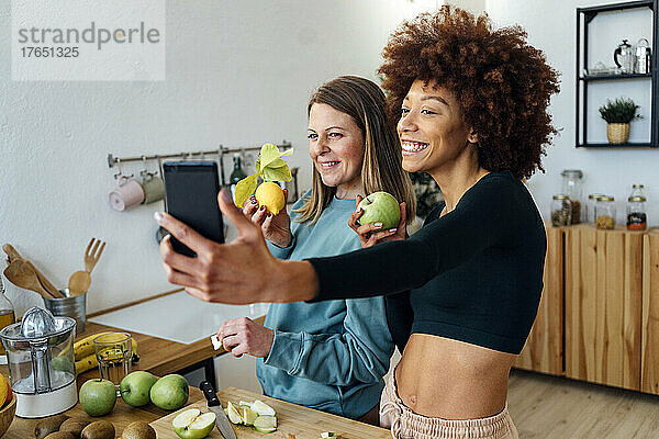 Glückliche Frauen mit frischen Früchten  die zu Hause in der Küche ein Selfie mit dem Smartphone machen