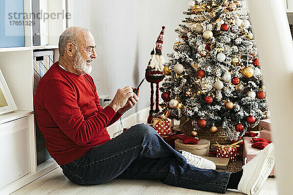 Glücklicher älterer Mann sitzt zu Hause am Weihnachtsbaum und nutzt sein Smartphone