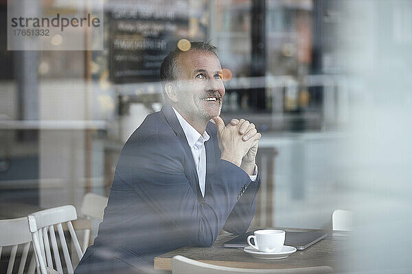 Lächelnder Geschäftsmann mit Kaffeetasse und Laptop  gesehen durch Glasfenster im Café
