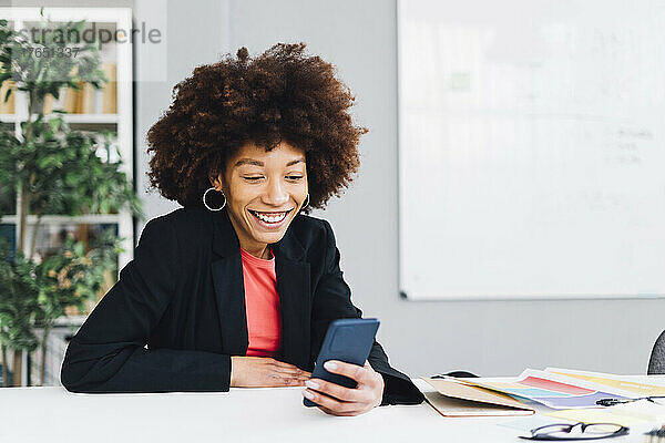 Glückliche Geschäftsfrau  die am Schreibtisch sitzt und im Büro ihr Mobiltelefon benutzt