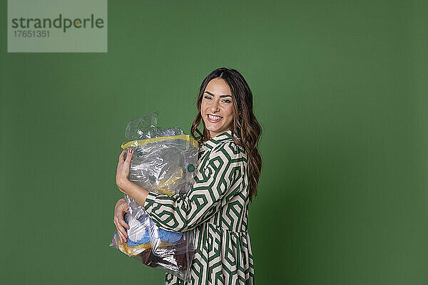 Glückliche junge Frau hält eine Tüte Plastikmüll vor grünem Hintergrund