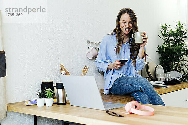 Lächelnder Freiberufler sitzt zu Hause mit Mobiltelefon auf der Küchentheke