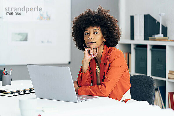 Nachdenkliche Geschäftsfrau mit der Hand am Kinn sitzt mit Laptop am Schreibtisch im Büro