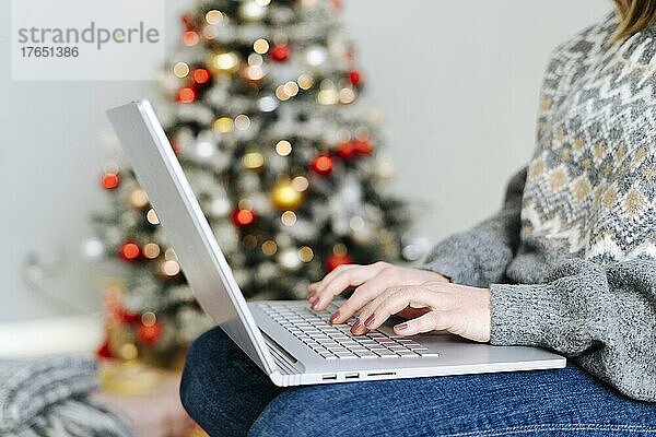 Hand einer Frau  die zu Hause in der Nähe des Weihnachtsbaums auf dem Laptop tippt