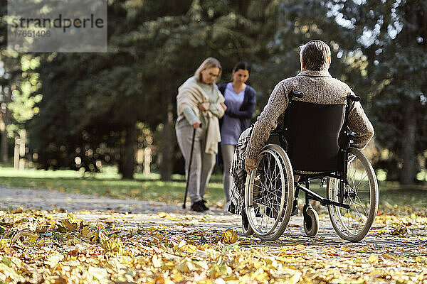 Älterer behinderter Mann sitzt im Rollstuhl vor einer Krankenschwester und geht mit einer Frau im Park spazieren