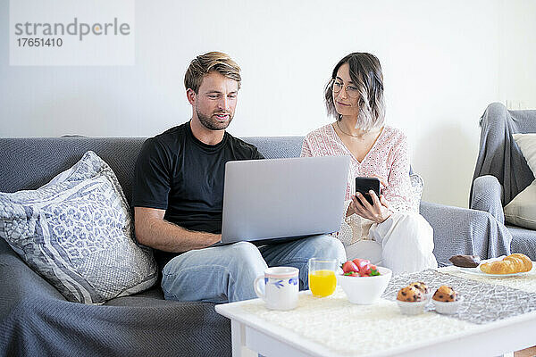 Mann benutzt Laptop und Frau hält Smartphone zu Hause auf dem Sofa