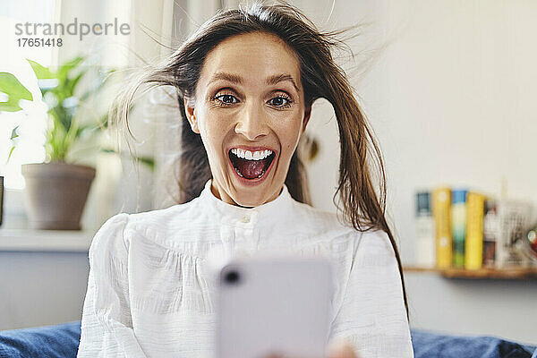 Überraschte Frau mit zerzausten Haaren  die zu Hause ihr Smartphone benutzt