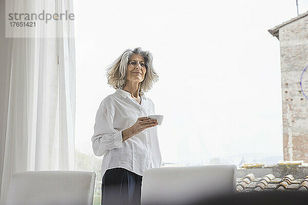 Lächelnde Frau mit Kaffeetasse  die zu Hause am hellen Fenster steht