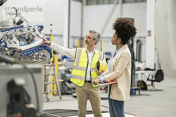 Ingenieur zeigt auf Roboterarm und diskutiert mit Geschäftsfrau  die Aktenordner in der Fabrik hält