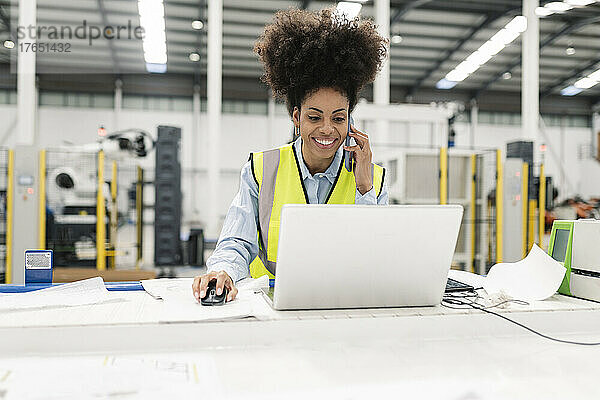 Lächelnder Ingenieur  der in der Fabrik am Laptop arbeitet und mit dem Mobiltelefon spricht