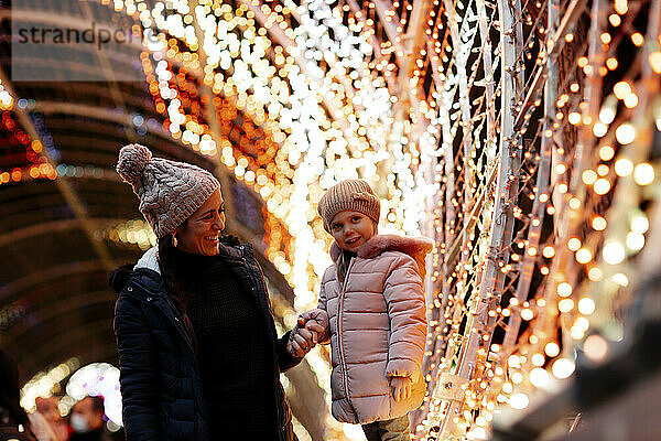 Happy woman looking at daughter standing by Christmas lights