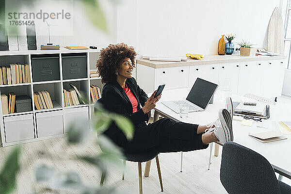 Junge Geschäftsfrau sitzt mit erhobenen Füßen auf dem Schreibtisch und hat ein Mobiltelefon im Büro