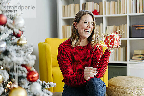 Fröhliche Frau mit Wunderkerze und Weihnachtsgeschenk sitzt zu Hause im Wohnzimmer