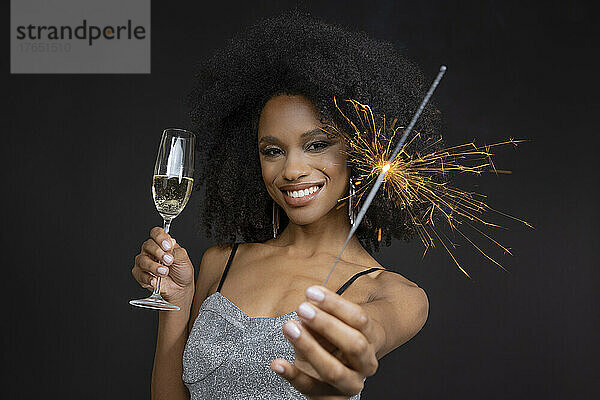 Lächelnde junge Frau mit Champagnerglas und Wunderkerze vor schwarzem Hintergrund