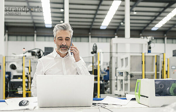 Geschäftsmann spricht mit Smartphone und steht mit Laptop am Schreibtisch in der Fabrik