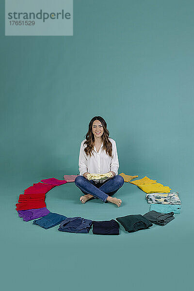 Smiling woman sitting cross-legged amidst clothes against blue background