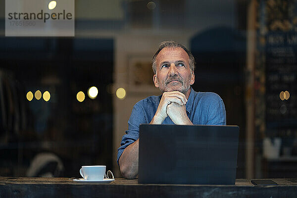 Nachdenklicher Geschäftsmann mit Laptop und Kaffeetasse sitzt am Tisch im Café