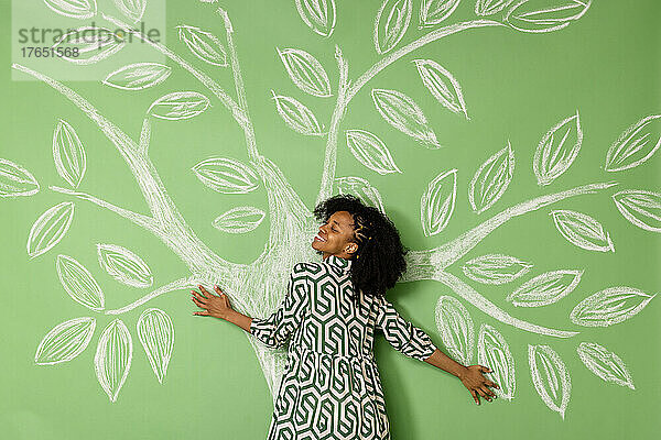 Lächelnde Frau mit geschlossenen Augen umarmt einen Baum  der eine grüne Wand zeichnet