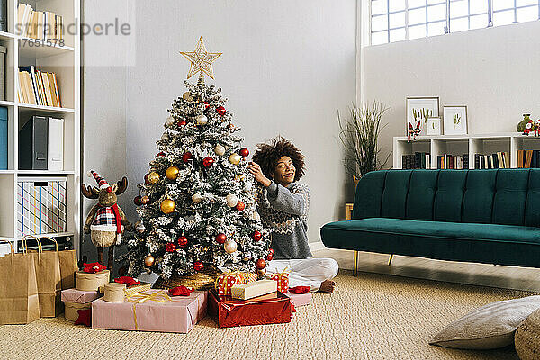 Lächelnde junge Frau sitzt mit gekreuzten Beinen und schmückt zu Hause im Wohnzimmer den Weihnachtsbaum