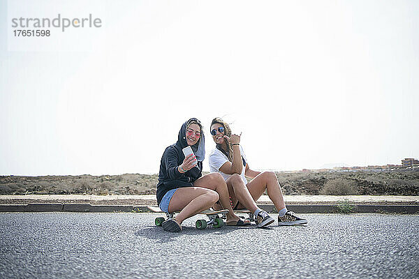 Freunde machen Selfies per Smartphone  indem sie an einem sonnigen Tag auf dem Skateboard sitzen