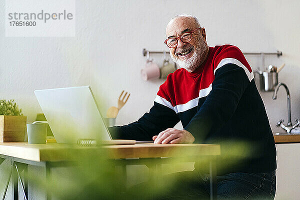 Glücklicher älterer Mann mit Brille sitzt zu Hause mit Laptop