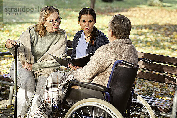 Ältere Frau und Krankenschwester mit Dokumenten betrachten behinderten Mann  der im Rollstuhl im Park sitzt
