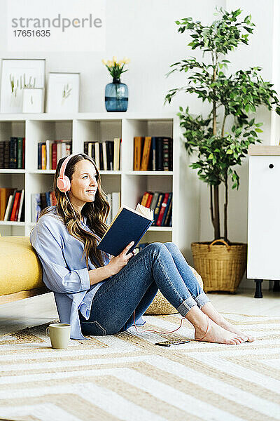 Lächelnde Frau mit Kopfhörern und Buch sitzt zu Hause auf dem Sofa auf dem Teppich