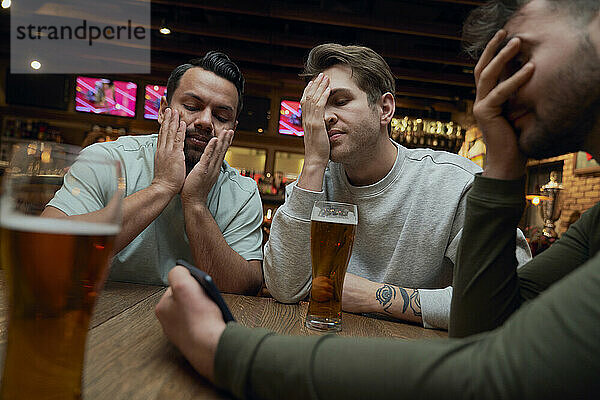 Drei frustrierte männliche Fußballfans trinken Bier und schauen sich ein Spiel auf dem Smartphone in einer Kneipe an