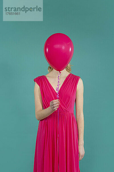 Young woman holding balloon in front of face standing against green background