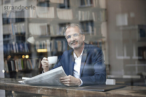 Lächelnder Geschäftsmann mit Kaffeetasse und Zeitung sitzt am Tisch im Café