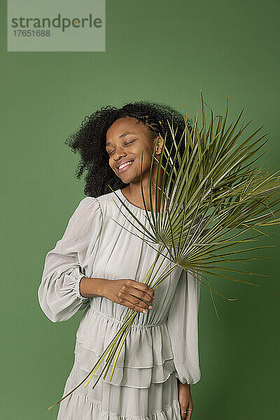 Lächelnde junge Frau mit geschlossenen Augen  die Palmzweige vor grünem Hintergrund hält