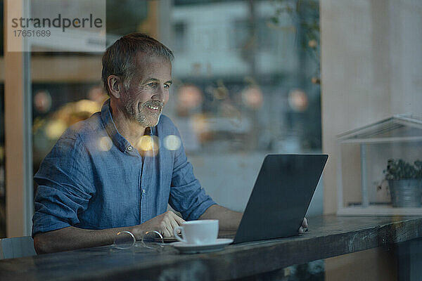 Lächelnder Geschäftsmann mit Laptop und Kaffeetasse sitzt am Tisch am Glasfenster im Café