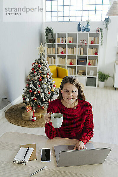 Lächelnde Frau mit Laptop und Kaffeetasse sitzt zu Hause im Wohnzimmer