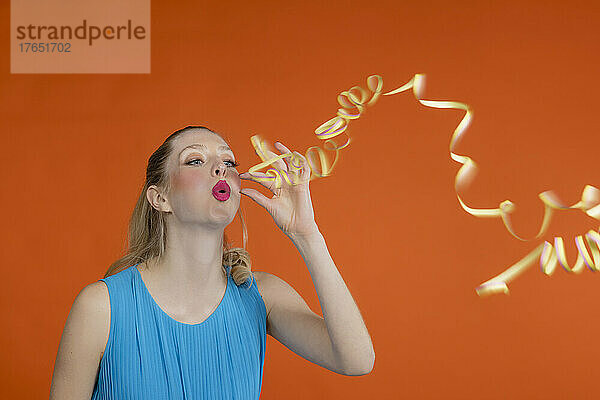 Playful young woman with blond hair blowing streamer against orange background