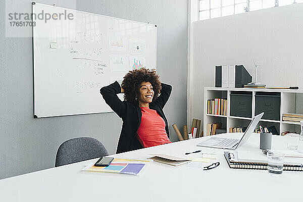 Lächelnde Geschäftsfrau mit den Händen hinter dem Kopf sitzt am Schreibtisch im Büro