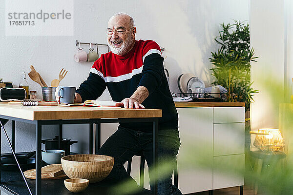 Fröhlicher älterer Mann mit Kaffeetasse sitzt am Tisch in der Küche