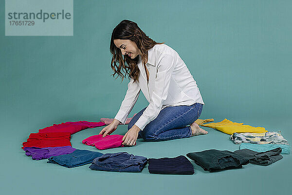 Smiling woman arranging t-shirt amidst clothes against blue background