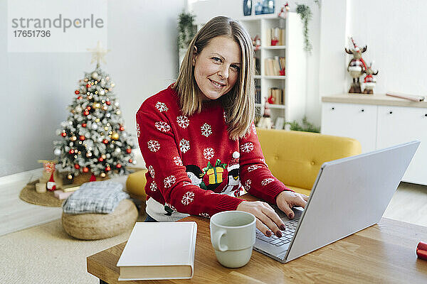 Lächelnde blonde Frau mit Laptop sitzt am Tisch im Wohnzimmer