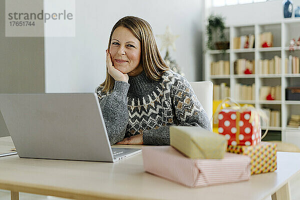 Glückliche Frau sitzt mit Laptop und Weihnachtsgeschenken zu Hause auf dem Tisch