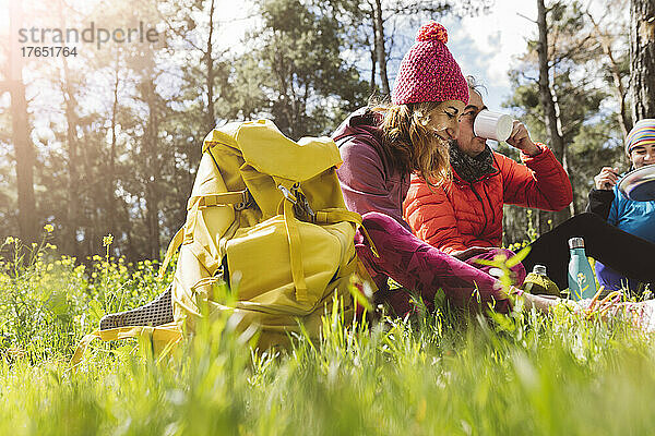 Glückliche Frau mit Rucksack  die an einem sonnigen Tag bei Freunden sitzt und im Wald isst und trinkt